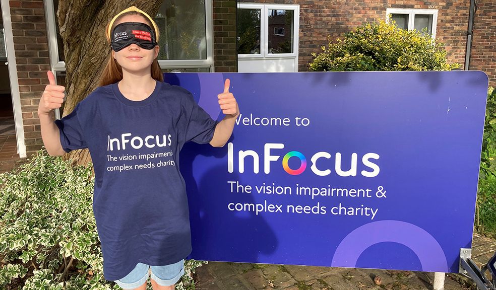 Photo is of an 11 year old girl, wearing a blind fold, standing in front of InFocus Charity Logo